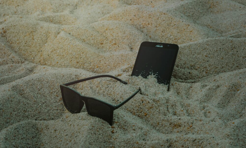 A smartphone and sunglasses partially buried in beach sand, capturing a summer vibe.