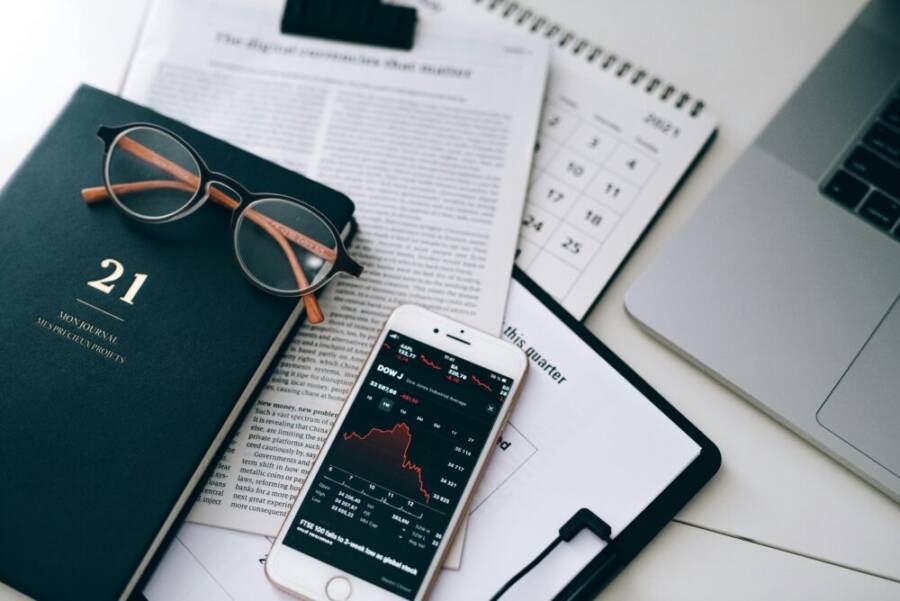 A stylish workspace featuring financial documents, eyeglasses, an iPhone displaying stock data, and a laptop.