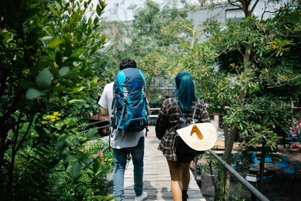 A couple with backpacks stroll on a wooden bridge surrounded by lush greenery, enjoying nature