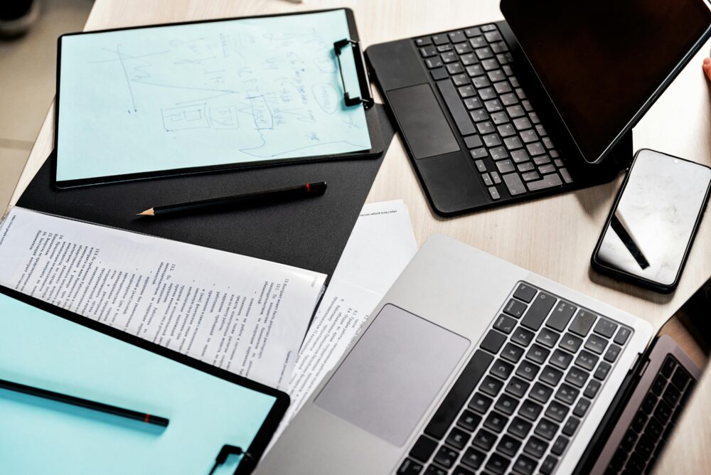 High-angle view of a modern workspace featuring laptops, tablet, clipboard, and documents.