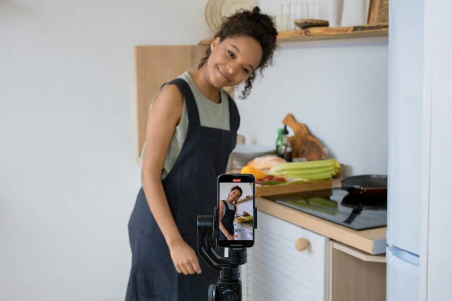 Smiling young woman filming a cooking video at home using a smartphone in her kitchen