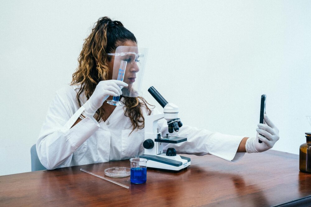 Female scientist in lab coat, using microscope and taking selfie, showcasing modern lab environment.