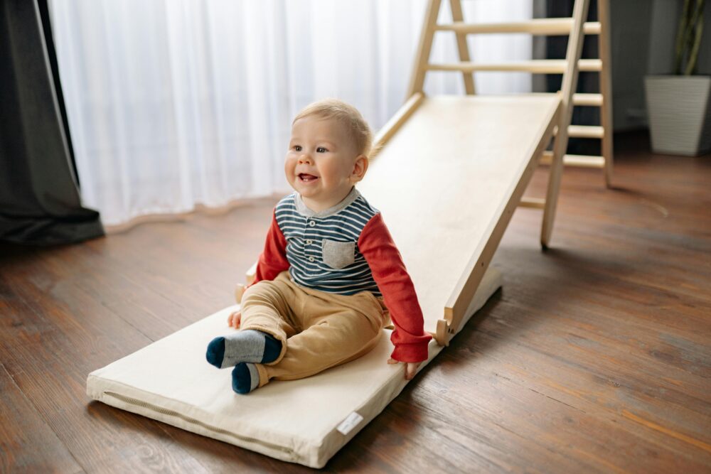 Adorable toddler smiling while sitting on a cushion at the bottom of an indoor wooden slide