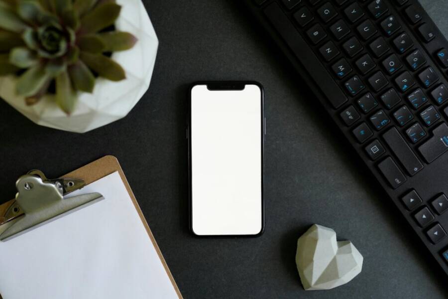 Top view of a smartphone, keyboard, succulent plant, and clipboard on a black desk.
