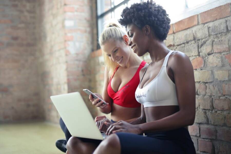 Two women in sportswear enjoy leisure time, sharing a laptop and smartphone in a gym setting against a brick wall