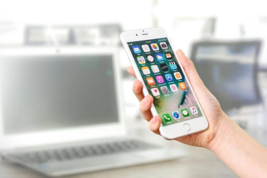 Close-up view of a smartphone displaying apps, held by a hand, with a blurred laptop in the background