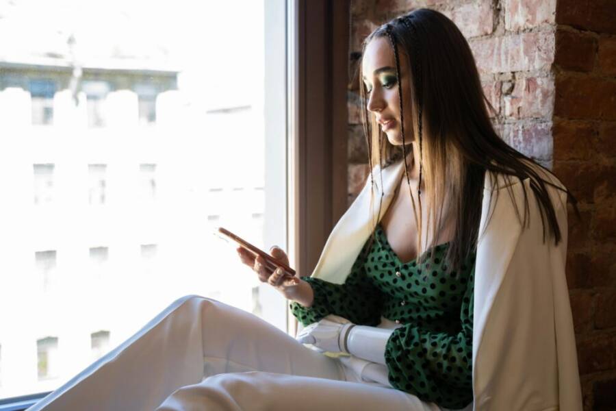 A woman with a prosthetic arm using a smartphone by the window indoors