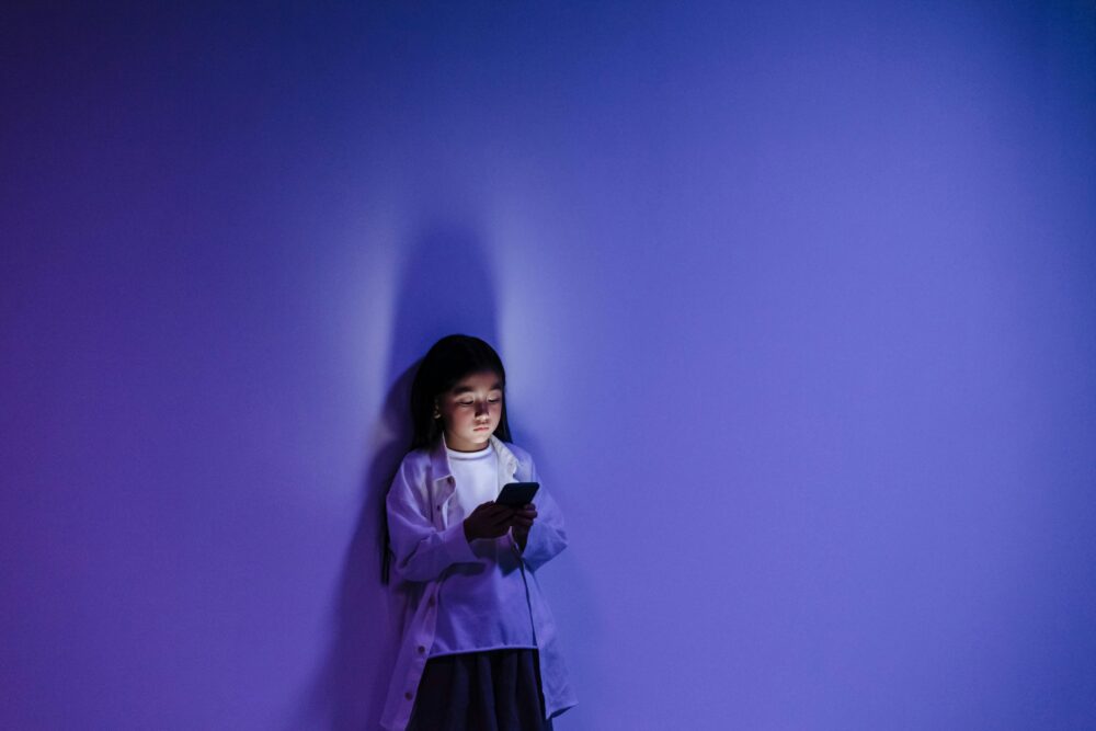 A girl standing against a blue-lit wall engrossed in her smartphone, creating a dramatic silhouette.