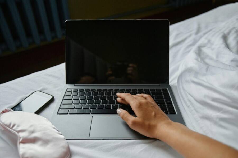 A hand resting on a laptop keyboard beside a phone and eye mask on a bed.