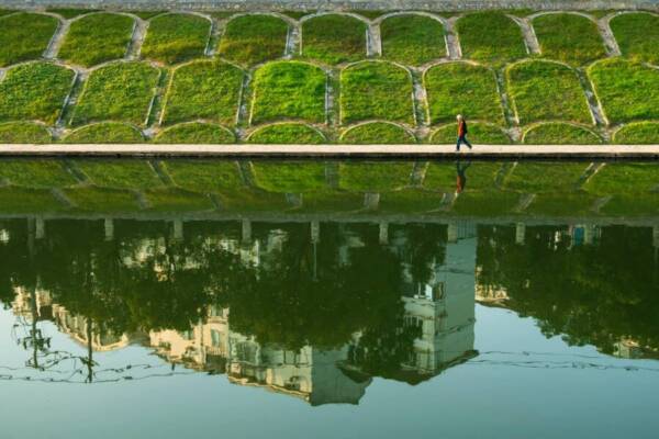 一個男子沿著河邊的小路行走，平靜的水面上倒映著城市的悲傷。