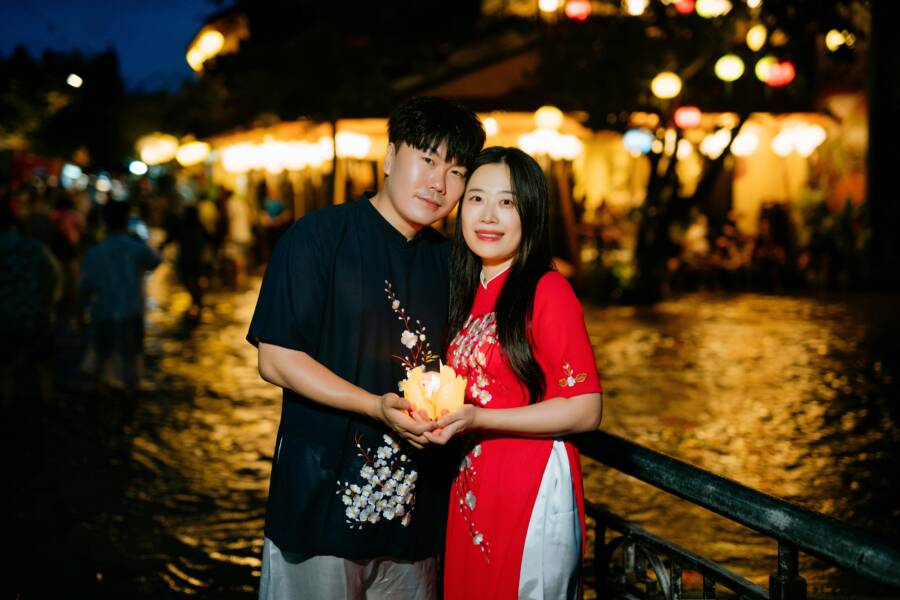 An Asian couple standing by a riverside, embracing and holding a lantern at night.