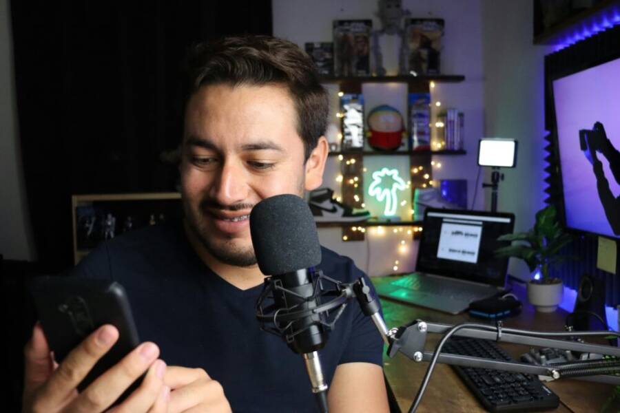 A man using a smartphone while seated at a desk with a microphone, surrounded by cozy decor