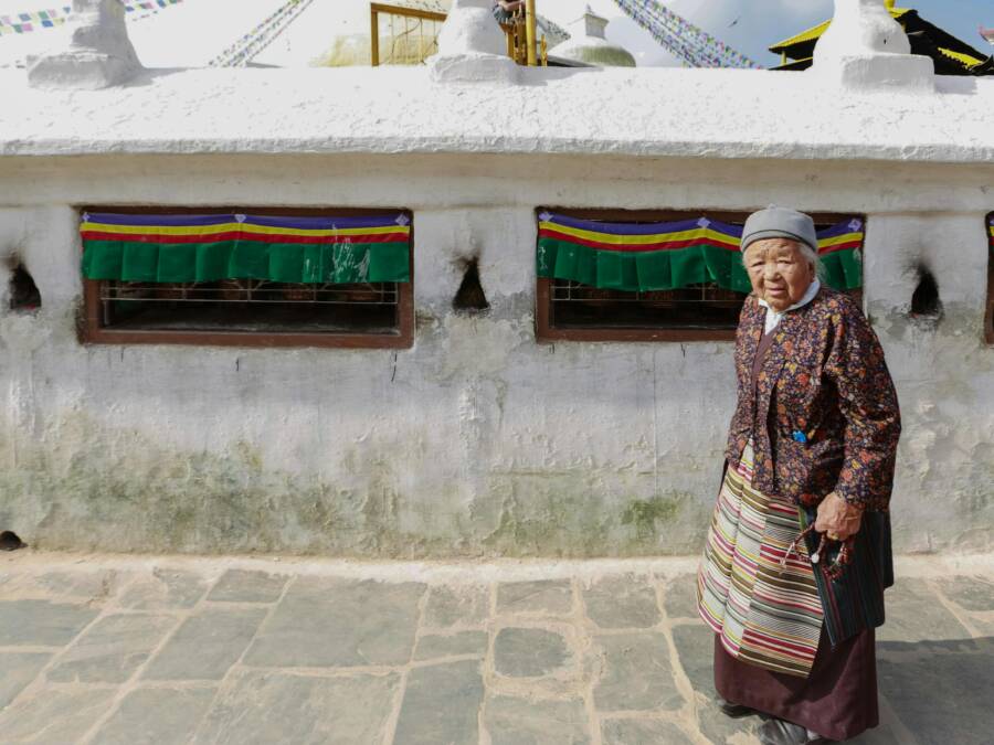 An elderly woman walks near a traditional Buddhist temple, wearing cultural attire