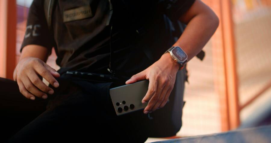 Casually dressed man with smartphone and smartwatch sitting outdoors, focusing on technology.