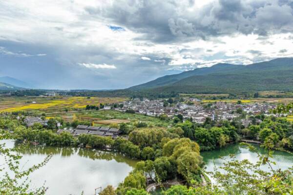 Picturesque view of a mountain town with lush fields and a serene lake under dramatic skies.