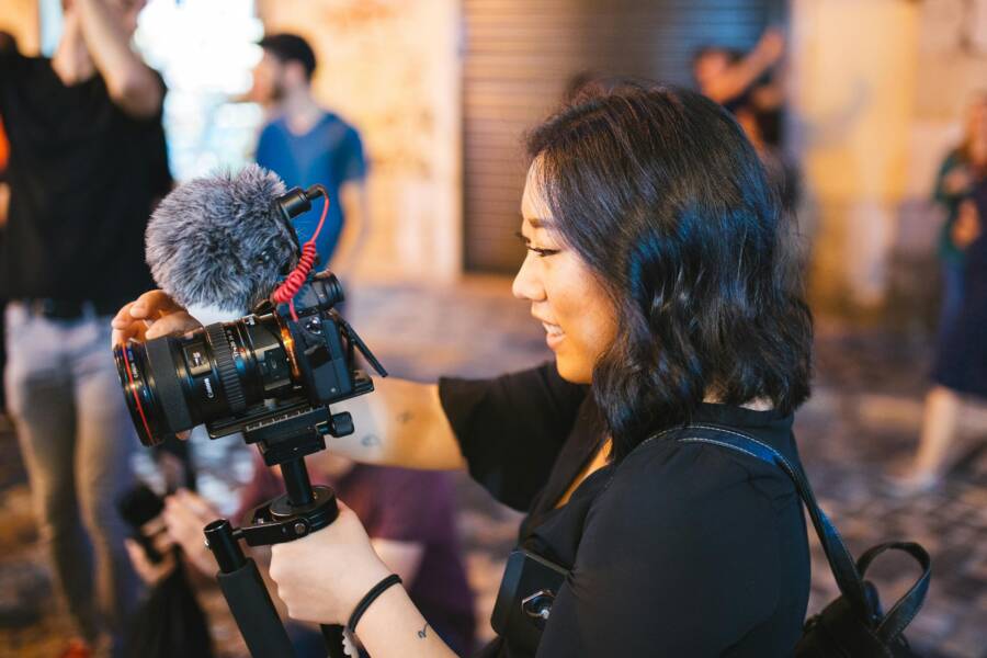 An Asian woman using a DSLR camera with a mic outdoors, engaged in filming or vlogging on a busy street