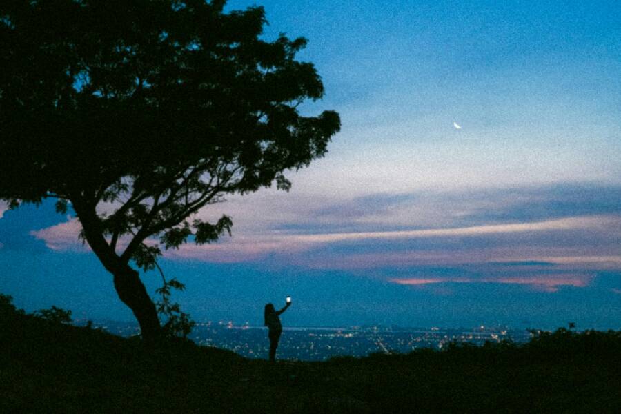 A silhouette of a person taking a photo against a cityscape during dusk with a visible crescent moon