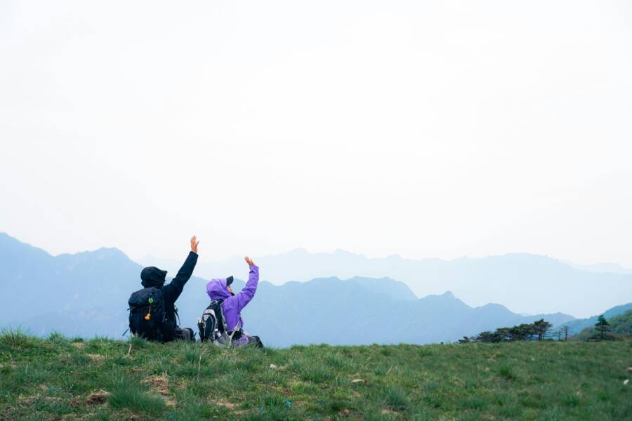 Two hikers wave enthusiastically atop a grassy hill with mountain views.