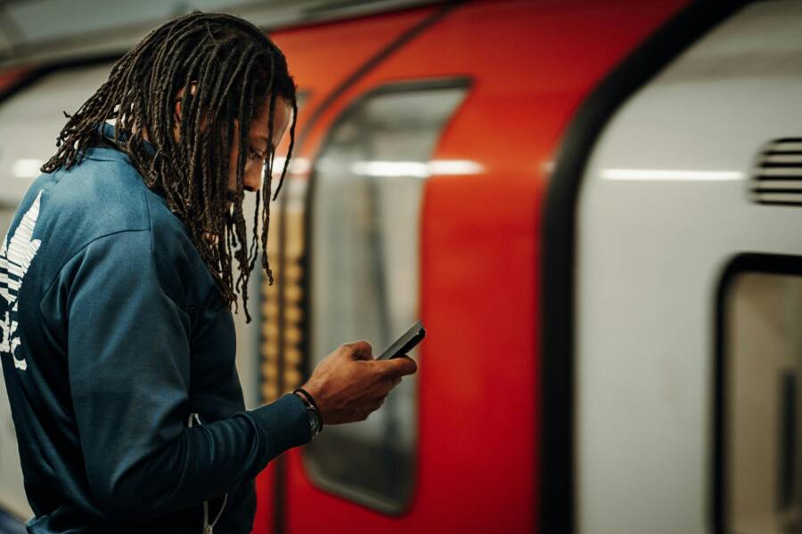 How to Speed Up Slow Hotspot Internet from Your Phone Young man with dreadlocks using smartphone on London Underground platform, waiting for train.