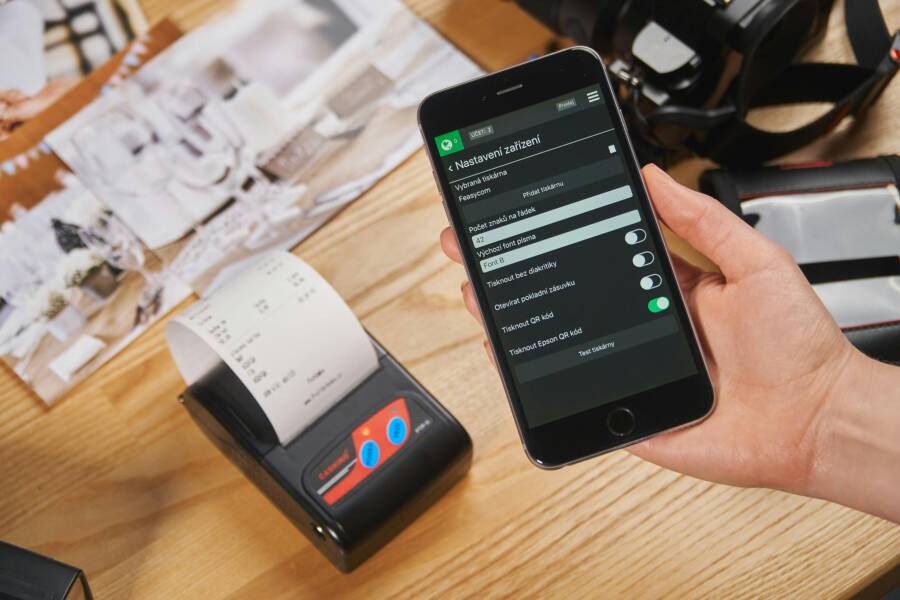 Close-up of a hand holding a smartphone with POS settings displayed, next to a receipt printer