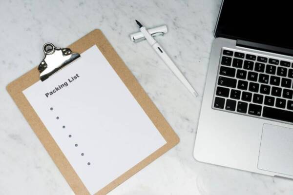 Flat lay of a packing list clipboard with a laptop and pen on a marble desk.
