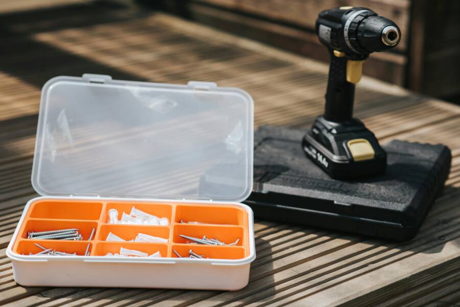 Cordless electric screwdriver placed on case on wooden surface near screw set with bolts and nails on street on blurred background