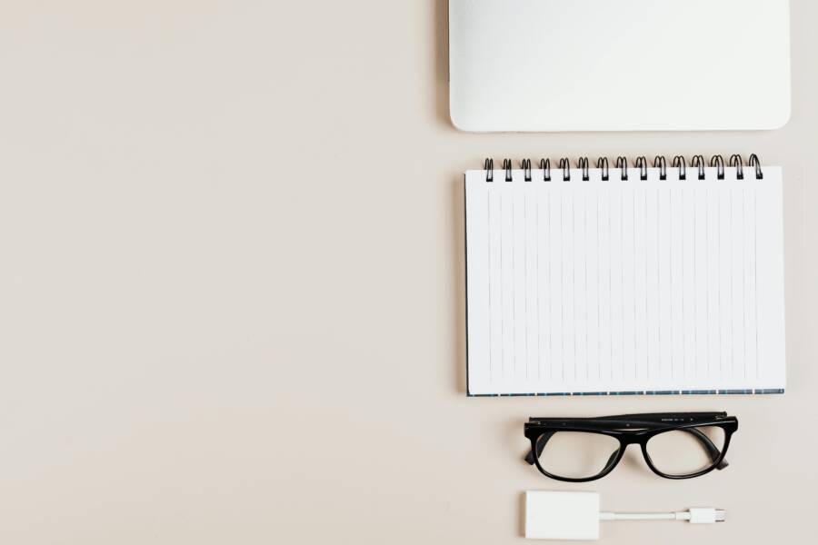 Top view of minimal office desk setup with notepad, laptop, glasses, and adapter.