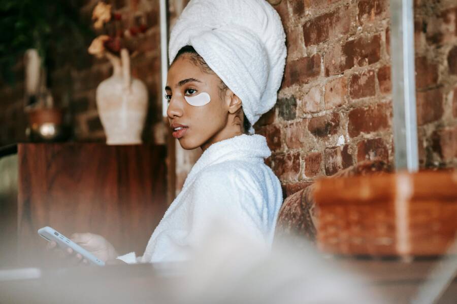 Side view of alluring African American female with eye patches in white bathrobe and towel on head messaging on mobile phone while resting at home