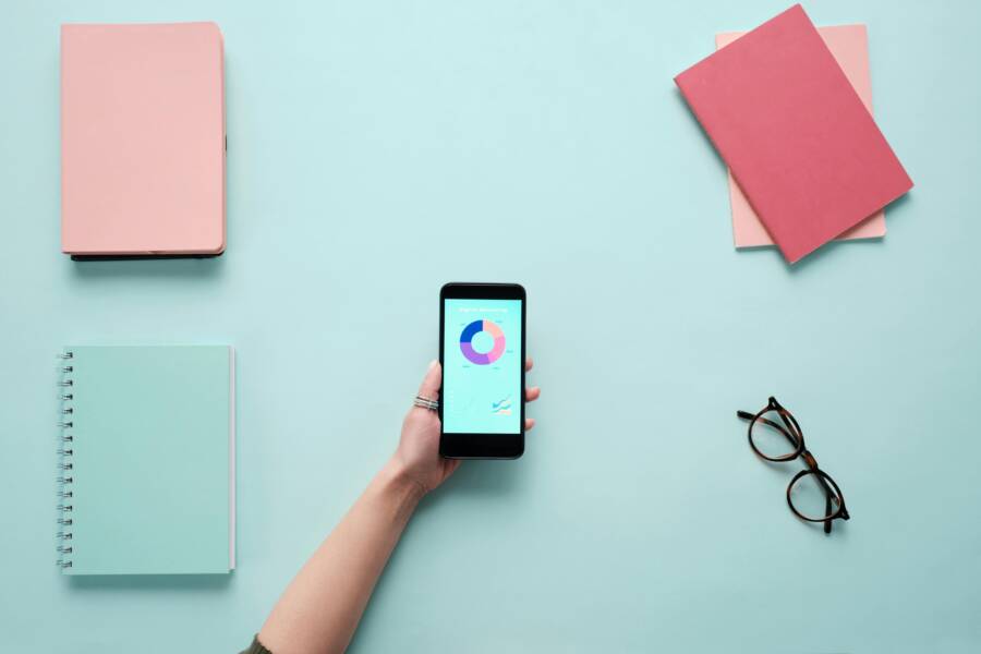 Minimalist flat lay with a smartphone displaying charts, surrounded by notebooks and glasses on a blue background