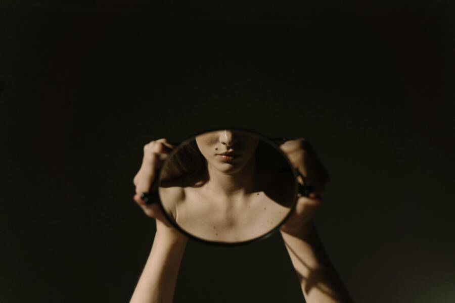 image: Artistic portrait of a woman holding a mirror, reflecting her face, with a dark, moody background