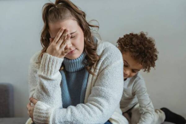 Frustrated mother with hand on forehead and closed eyes sitting near African American son near wall in room at home