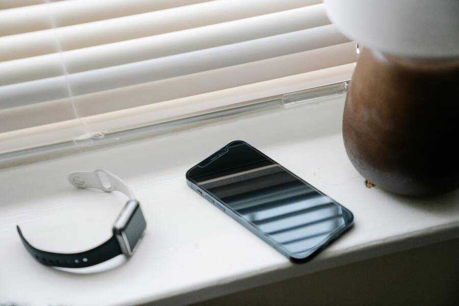 A smartphone and smartwatch placed on a windowsill with morning light filtering through blinds.