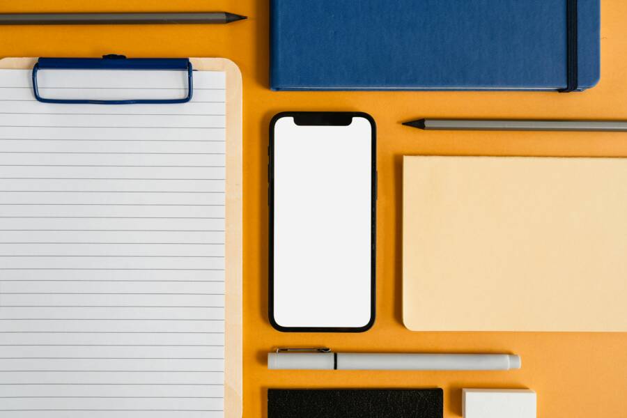 A top view of office essentials including a smartphone, clipboard, and stationery on a yellow surface