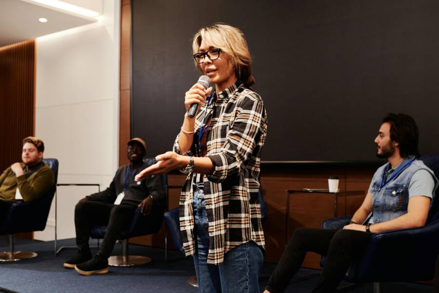A confident woman speaks into a microphone during a panel discussion indoors.