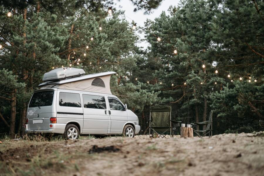 A campervan set up in a serene forest campsite adorned with hanging lights. Tranquil outdoor escape.